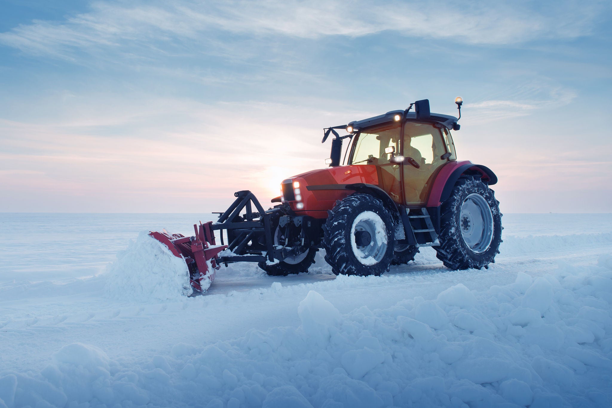 A red tractor navigates through a snow-dusted field, preparing the land for planting under a bright blue sky.