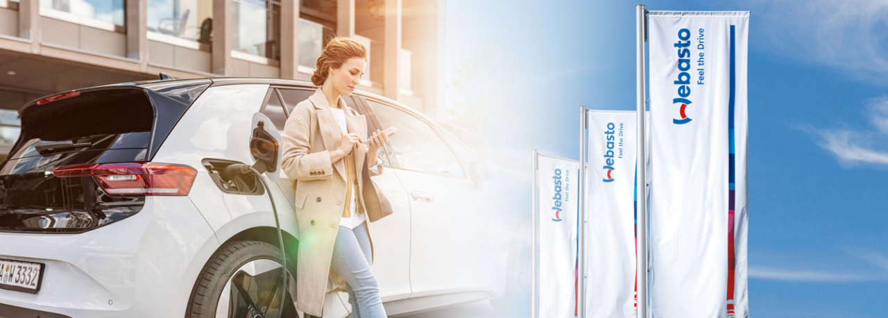 Frau lädt ein Elektrofahrzeug an einer Ladesäule vor Webasto-Fahnen, symbolisiert Webasto als systemischen Partner für Ladeinfrastruktur, Investor Relations.