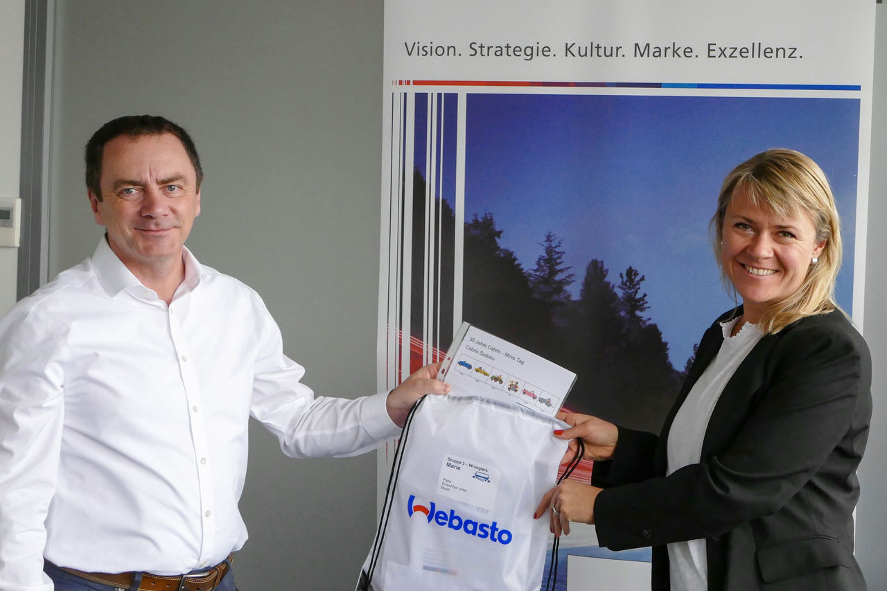 Webasto Careers (DE) employer-branding: colleagues exchanging a branded welcome kit by a 'Vision. Strategie. Kultur' backdrop, showing Webasto as system partner.