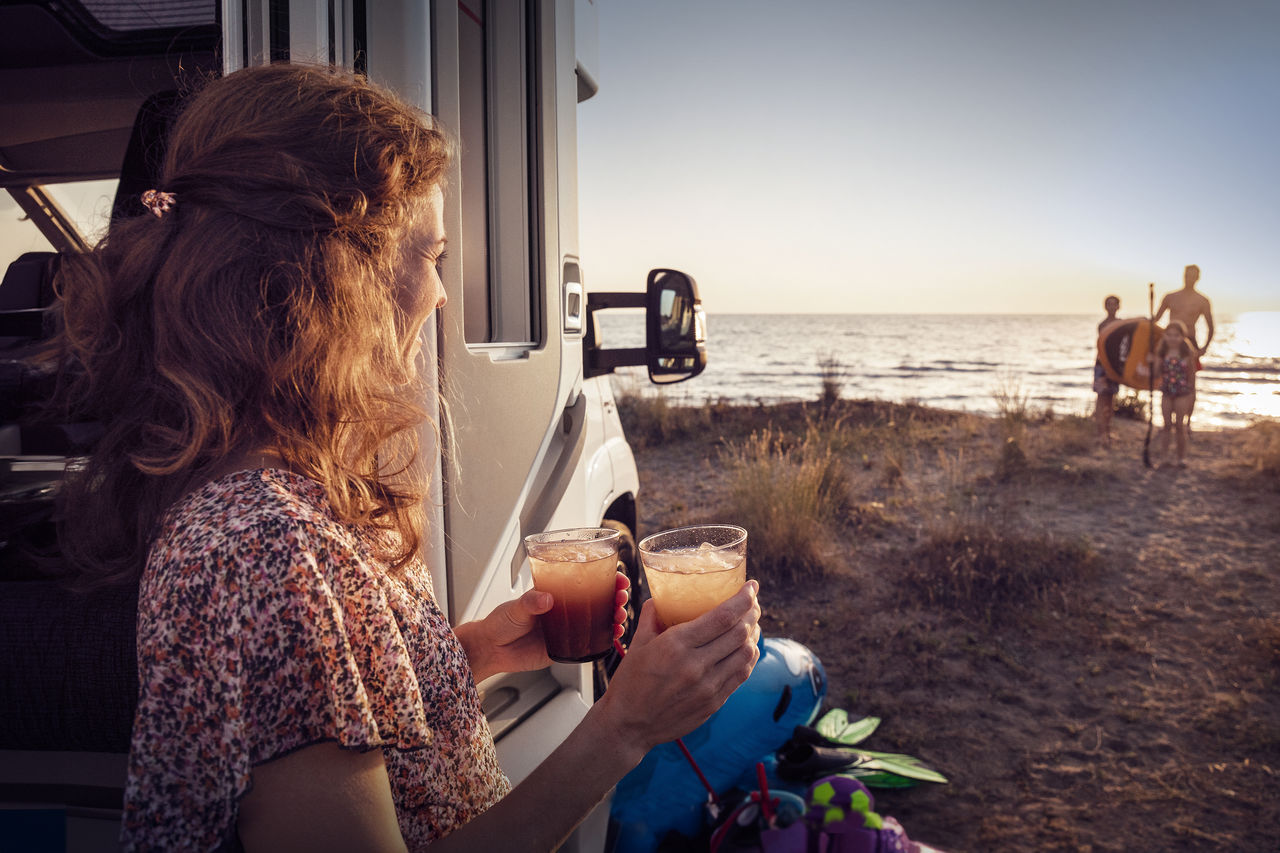 Woman holding two drinks beside a motorhome at the beach at sunset, highlighting Webasto roof systems, thermal management and auxiliary power solutions for leisure travel.